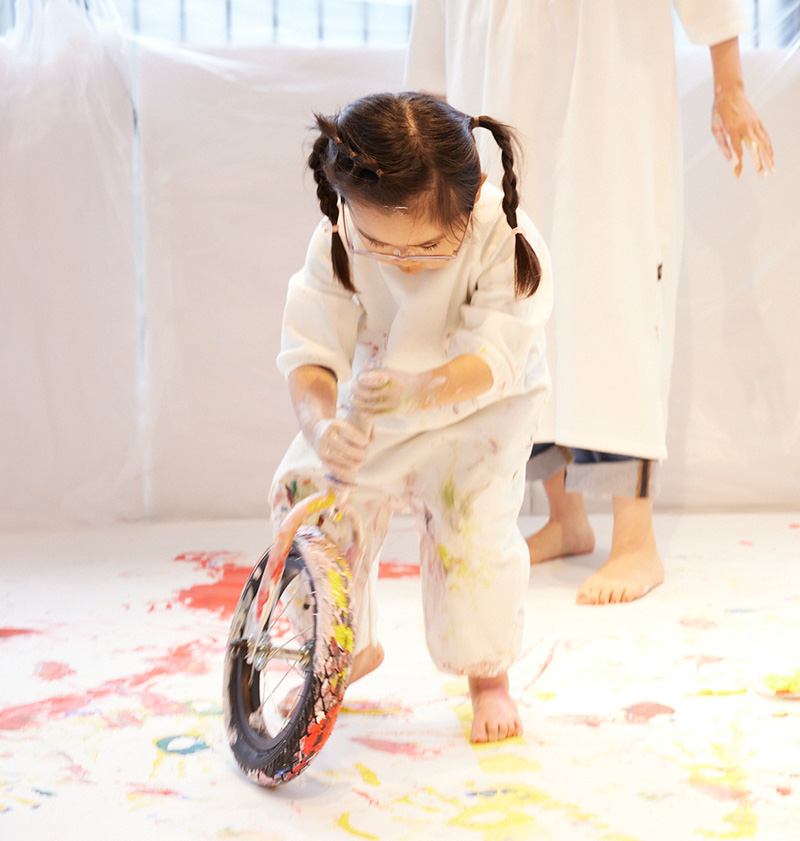 かぞくのおえかきウェアを着てへんしんバイクのタイヤを転がしてえのぐ遊びを楽しんでいる子供